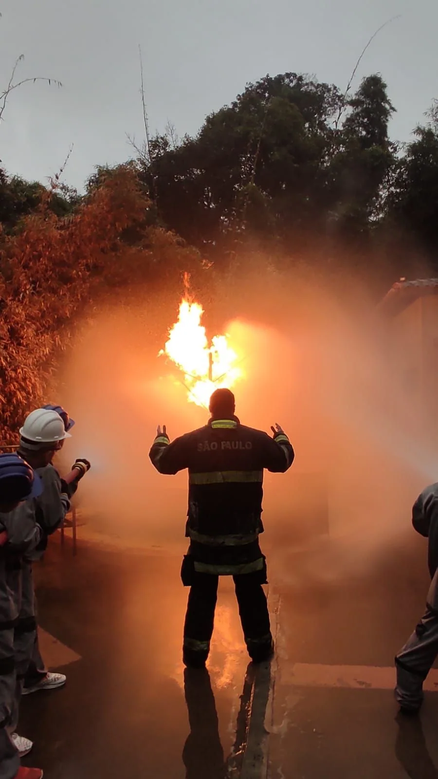 Instrutor conduzindo exercício de combate a incêndio diante de chama de grande porte, com a turma posicionada ao redor